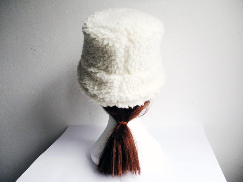 Puede incluir: Un gorro blanco y peludo de estilo bucket con una coleta marr&oacute;n que sobresale por debajo.