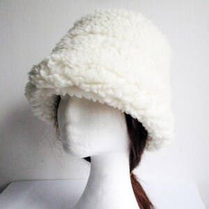 Puede incluir: Un gorro blanco y esponjoso de estilo bucket hecho de un material suave y esponjoso. El gorro se lleva sobre una cabeza de maniqu&iacute; blanca con pelo marr&oacute;n.