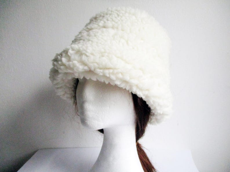 Puede incluir: Un gorro blanco y esponjoso de estilo bucket hecho de un material suave y esponjoso. El gorro se lleva sobre una cabeza de maniqu&iacute; blanca con pelo marr&oacute;n.