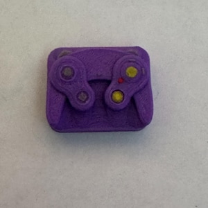 May include: A purple 3D printed object shaped like a video game controller. The controller features two circular joystick areas, a small red button, and two yellow circular buttons set into the textured purple plastic surface.