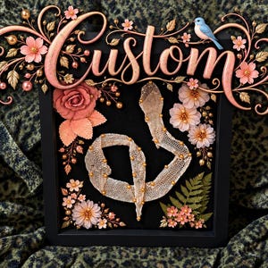 May include: A black shadow box frame containing a snake skin, decorated with pink fabric flowers, gold beads, and a blue bird figurine. The word Custom is written in large, ornate, metallic pink script above the frame with floral accents.