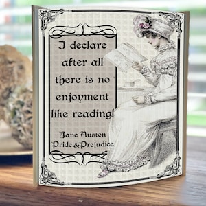 May include: A decorative wooden plaque featuring a quote from Jane Austen's Pride & Prejudice. The quote reads "I declare after all there is no enjoyment like reading!" and is surrounded by a black and white ornate frame. The plaque features a vintage illustration of a woman reading a book.