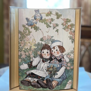 Puede incluir: Una impresión enmarcada de estilo vintage con dos muñecas de trapo sentadas bajo un arco floral. Las muñecas están vestidas con ropa colorida y sostienen un ramo de flores. La impresión tiene un marco marrón claro.