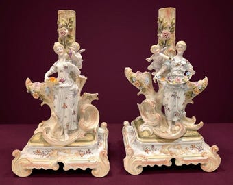 May include: A pair of porcelain candle holders featuring sculpted figures of people and cherubs amidst floral motifs. The bases are ornate with scrollwork and painted floral details in pastel tones of pink, green, and gold.