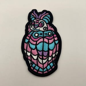 May include: Embroidered patch featuring a stylized creature design. The patch showcases a vibrant color palette of pink, blue, purple, and white, outlined with black stitching. The design has a unique, whimsical aesthetic, suitable for clothing or accessories.