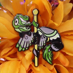 May include: A detailed enamel pin featuring a carousel lizard design. The pin has a black outline with green, gray, and yellow accents. The lizard is mounted on a yellow pole with a green leaf design. The pin is set against a yellow flower.