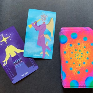 Puede incluir: Dos cartas de tarot, La Estrella y El Juicio, con una baraja de cartas en el fondo. La carta de La Estrella es morada con una figura amarilla y una estrella en el cielo. La carta de El Juicio es azul con una figura morada con alas y una trompeta. La baraja de cartas es rosa y azul con un patr&oacute;n circular.