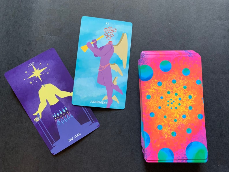 Puede incluir: Dos cartas de tarot, La Estrella y El Juicio, con una baraja de cartas en el fondo. La carta de La Estrella es morada con una figura amarilla y una estrella en el cielo. La carta de El Juicio es azul con una figura morada con alas y una trompeta. La baraja de cartas es rosa y azul con un patr&oacute;n circular.
