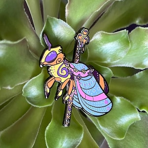 May include: A yellow and purple enamel pin of a bee wearing a floral crown and a blue and pink patterned dress. The bee is holding a staff with a honeycomb on top.