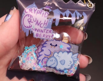 Puede incluir: Un amuleto de bolsa de plástico transparente con un diseño morado y blanco. La bolsa tiene un diseño morado y blanco con el texto "Cryptid Crunch...Winter Berry!" y un monstruo morado y blanco en su interior. La bolsa está llena de purpurina.