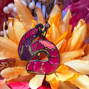 May include: A red and purple enamel pin of a cartoon worm with a crown on its head, holding a staff with a star on top. The worm is wearing a pink and yellow patterned dress. The pin is set against a background of orange and yellow fabric.