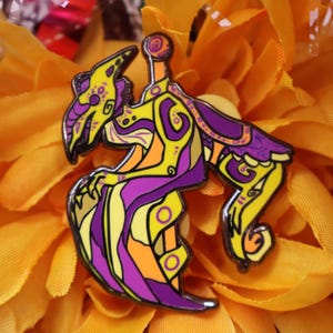 Puede incluir: Un colorido pin de esmalte con un diseño estilizado y abstracto de criatura. El pin tiene una combinación de colores amarillo, morado y naranja con contornos negros. El pin está sobre un fondo floral amarillo brillante.