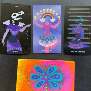Puede incluir: Tres cartas de tarot con fondos negros e ilustraciones coloridas. Las cartas se titulan "Muerte", "Nueve de Copas" y "Nueve de Espadas".