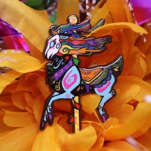 May include: A colorful enamel pin featuring a carousel deer design. The deer has a white face, black body, and is adorned with vibrant blue, pink, orange, and purple accents. The pin is set against a backdrop of yellow.
