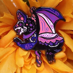 Könnte beinhalten: Dekorative Emaille-Anstecknadel mit einer stilisierten Fledermaus im Stil des Día de los Muertos. Die Fledermaus ist schwarz mit lila, rosa und weißen Akzenten. Die Flügel sind rosa und lila mit weißen Details.