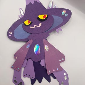 May include: A layered papercraft figure in shades of purple, featuring a large hat, yellow eyes with red pupils, and a small, smiling mouth. The figure has a central iridescent gem and iridescent accents.