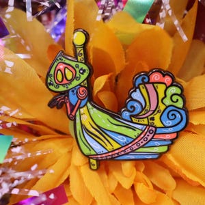 May include: A colorful enamel pin featuring a stylized, whimsical design. The pin showcases a vibrant palette of green, yellow, blue, and orange, with intricate details and a black outline. The pin is set against a backdrop of yellow petals.