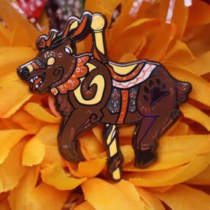 May include: A brown and yellow enamel pin in the shape of a carousel bear. The bear has a detailed design with orange and yellow accents, a paw print, and a yellow pole. The pin is set against a background of yellow fabric.