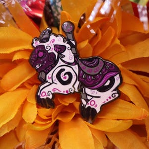 May include: A decorative enamel pin in the shape of a carousel horse. The pin features a white base with purple and black accents, including a detailed saddle and face design. The pin is set against a bright yellow flower.