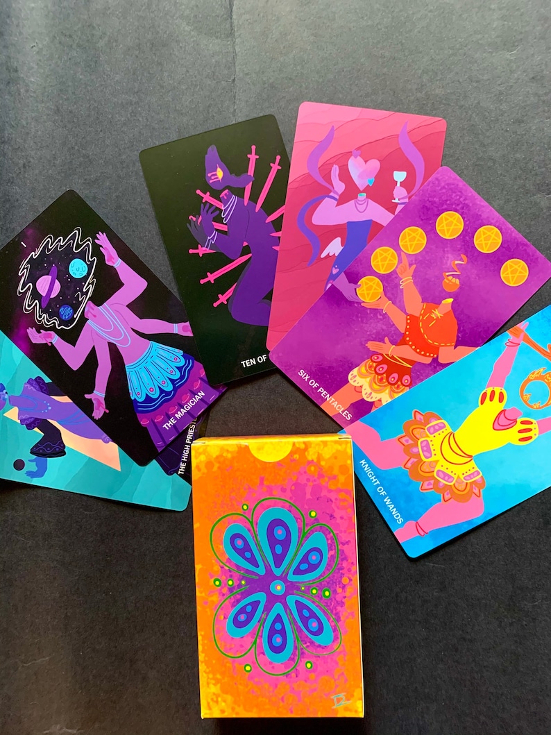 Puede incluir: Un juego de cartas de tarot con ilustraciones coloridas de personas y s&iacute;mbolos. Las cartas est&aacute;n dispuestas en c&iacute;rculo alrededor de una caja con un dise&ntilde;o floral. Las cartas incluyen "El Mago", "Diez de Espadas", "Seis de Pent&aacute;culos" y "Caballero de Varitas".
