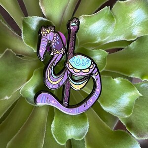 May include: Purple and blue enamel pin of a flamingo on a carousel pole. The flamingo has a pink and yellow saddle and is decorated with flowers.