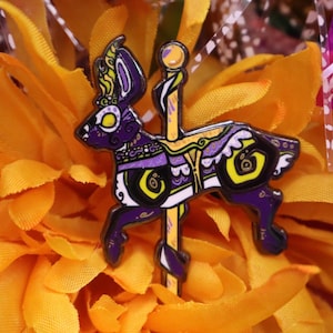 May include: A purple and yellow carousel-style pin featuring a stylized rabbit design. The pin has intricate details with black, white, and green accents. The pin is mounted on a yellow pole with a gold ball at the top. The background is a bright orange.