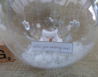 Op de afbeelding: Een glazen sneeuwbol met een wit sneeuwpopfiguurtje erin. De sneeuwpop houdt een bord vast met de tekst "Will you marry me?" Er hangen kleine witte sneeuwvlokken aan de bovenkant van de bol.