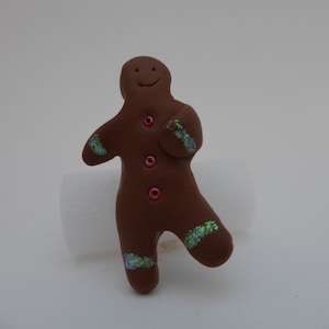 May include: A brown gingerbread man figurine with a smiling face and red buttons. The figurine has green glitter accents on its arms and legs.