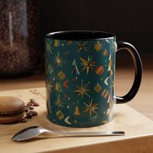 May include: A ceramic mug with a black handle and interior, featuring a teal exterior decorated with a repeating pattern of multi-coloured starbursts, striped spherical ornaments, geometric trees, and crescent shapes in shades of gold, orange, and white.