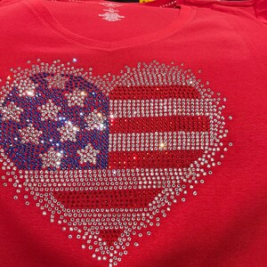 K&ouml;nnte beinhalten: Rotes T-Shirt mit einem herzf&ouml;rmigen Design aus Strasssteinen. Das Design ist eine amerikanische Flagge mit roten, wei&szlig;en und blauen Strasssteinen.