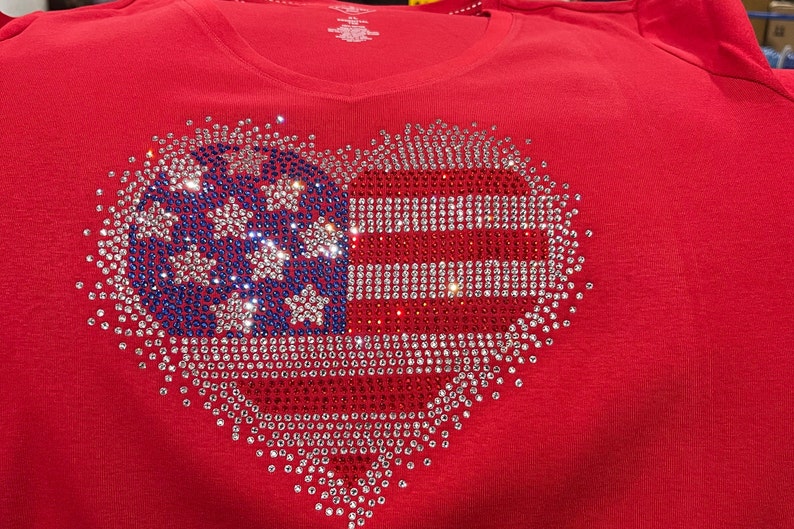 K&ouml;nnte beinhalten: Rotes T-Shirt mit einem herzf&ouml;rmigen Design aus Strasssteinen. Das Design ist eine amerikanische Flagge mit roten, wei&szlig;en und blauen Strasssteinen.