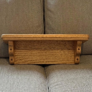 Puede incluir: Un estante de pared rectangular de madera con un acabado de grano natural en tonos marrón claro. El estante incluye una superficie superior plana y está sostenido por dos soportes de madera tallada a los lados, cada uno con dos detalles oscuros.