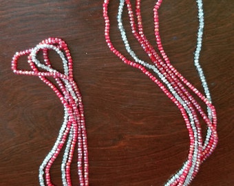 May include: A multi-strand necklace featuring alternating strands of red and silver-colored beads. The necklace has a silver clasp and is displayed on a dark brown wooden surface. The beads appear to be small and faceted.