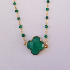 May include: A gold-colored necklace with a green clover-shaped pendant. The necklace features small green beads strung along a gold chain. The clover pendant is a faceted, vibrant green color, set in a gold frame.