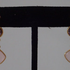 May include: A pair of gold-toned earrings with a post and a dangling design. Each earring features a round, glittery purple stone and a light pink, square-shaped stone. The earrings are displayed on a black stand.