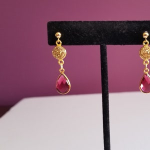 May include: Gold-toned dangle earrings with a round, textured top and a teardrop-shaped, deep pink gemstone. The earrings are displayed on a black stand against a purple background. The earrings have a classic and elegant design.