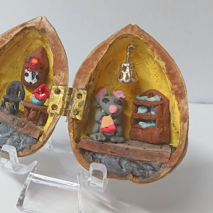 May include: A miniature house made from two halves of a walnut shell. The house is decorated with a small chair, a table, a lamp, and a tiny mouse figure. The house is open and the interior is painted yellow.