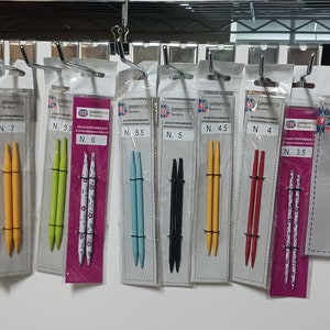 May include: A variety of knitting needles in different sizes and colors are displayed on a white background. The needles are packaged in clear plastic bags with labels indicating the size of the needles. The labels also include the brand name "DAMARAPITCH" and the text "Double Pointed".