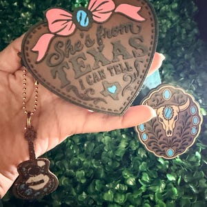 May include: A heart-shaped brown leather-like keychain with a pink bow and the text She's from TEXAS I CAN TELL, featuring a small Texas-shaped cutout. Attached is a guitar-shaped charm and a separate round charm with a steer skull design.