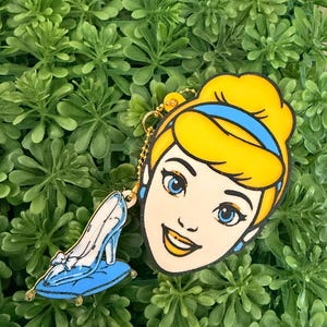 May include: A keychain featuring a flat, cartoon-style illustration of a character with blonde hair and a blue headband, attached to a smaller charm of a light blue high-heeled shoe resting on a blue pillow, connected by a gold ball chain.