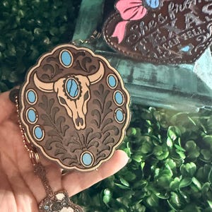 May include: A round brown leather keychain featuring an embossed steer skull with turquoise accents and a small guitar-shaped charm. A second heart-shaped leather piece displays the text "She's from TEXAS I CAN TELL" with a pink bow.