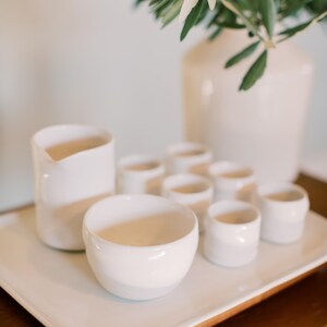 May include: A white ceramic set consisting of a small pitcher, a wide bowl, and six small cups arranged on a rectangular white tray. The pieces have a smooth, glossy finish and a simple, rounded shape, creating a cohesive collection for serving.
