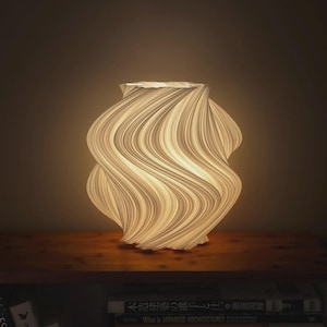 May include: A white, cylindrical table lamp with a spiraling, wavy, ribbed texture that emits a warm, soft glow. The lamp features a complex, layered design that creates deep shadows and highlights along its vertical, undulating surface.