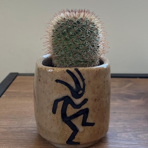 May include: A small, cylindrical, speckled tan ceramic planter featuring a black hand-painted figure of a dancing person. A round, green, spiky cactus with reddish-brown spines is planted inside the pot, extending above the rim.