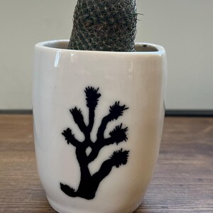 Puede incluir: Una pequeña maceta cilíndrica de cerámica blanca con una silueta negra de un árbol de Josué en la parte frontal. La maceta contiene un cactus verde, redondo y espinoso que sobresale por la abertura superior.