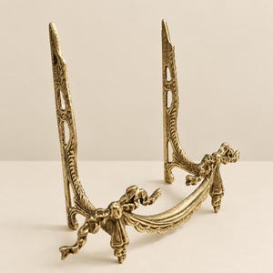 May include: A gold-toned metal display stand featuring two vertical uprights with intricate embossed detailing and a curved front base adorned with sculpted ribbon and bow motifs. The stand is designed to hold items upright.