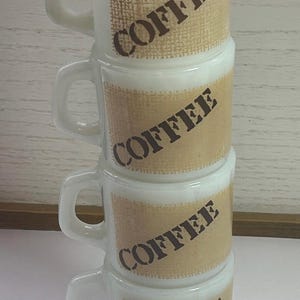 May include: A stack of four white coffee mugs with a brown burlap-style band featuring the word "COFFEE" in a dark brown, angled font. Each mug has a handle and a slightly flared rim.