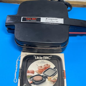 May include: A black "Little MAC" fast cooker with a handle and power cord. The appliance has a rectangular shape with a hinged top. A brochure with the same branding is below the cooker, showing images of cooked food.