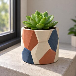 Puede incluir: Una maceta cilíndrica con un patrón de superficie geométrico y facetado en tonos alternos de naranja terracota, azul marino y crema. Una planta suculenta de color verde vibrante está plantada dentro del recipiente.