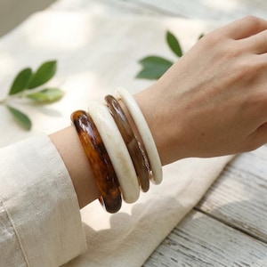May include: A set of four stacked bangle bracelets featuring a mix of smooth textures and colors. The collection includes one dark brown tortoiseshell pattern, one solid cream, one translucent brown, and one solid white bangle bracelet.
